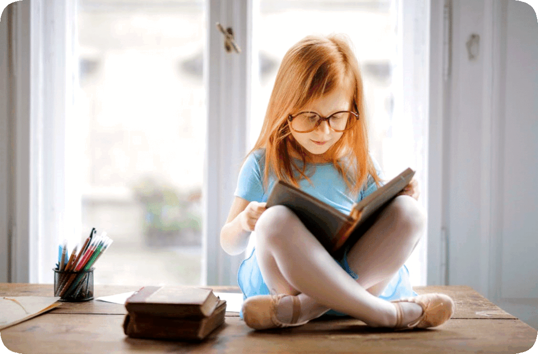 Picture of a girl reading a book while sitting on a table next to a window.