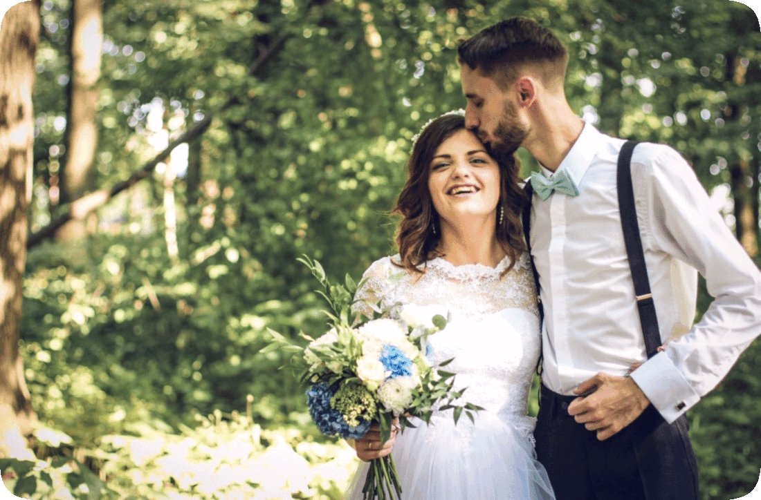 Picture of a happy smiling wedding couple standing outside in a wooded area, with the bride holding a bouquet of flowers and the groom kissing the bride’s forehead.