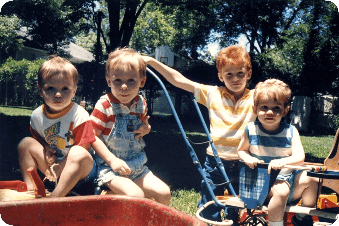 Picture of four boys playing outside in a grassy yard on a sunny day under the shade of a tree.