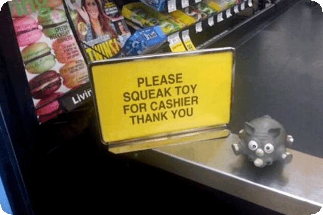 Picture taken inside a shop at the cashier station with a sign reading, ‘Please Squeak Toy For Cashier, Thank You’ and a gray toy plastic elephant squeaky toy next to it.