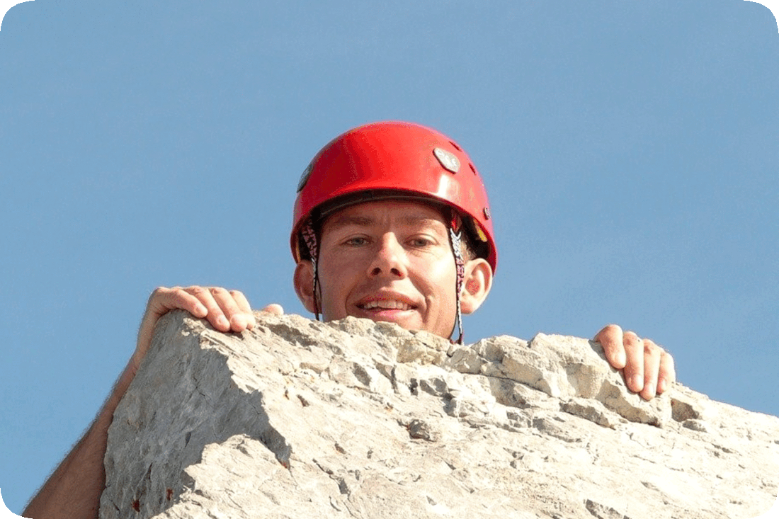 Picture of a man looking over the top of a large rock he is climbing, with a clear blue sky in the background.