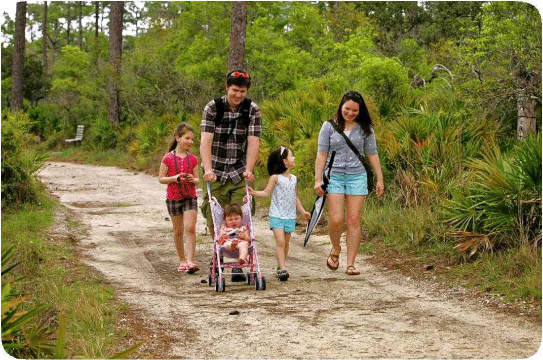 Picture of a family of 5, with a husband, wife, and 3 daughters, strolling through a park on a sandy trail, with green grass and semi-tropical green leafy plants and trees on both sides of the trail.