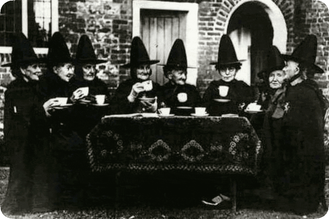 A very old black and white photograph of eight Welsh women dressed in black dresses and pointed black hats, seated at a table and holding cups of tea and saucers.