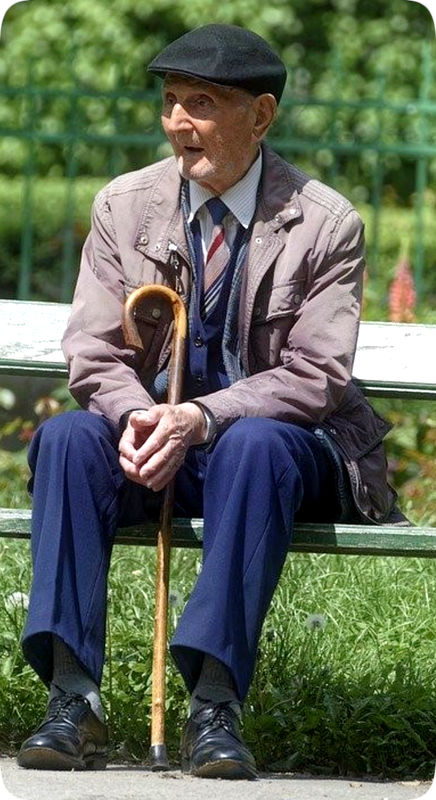 Picture of a man sitting on a wooden bench with his walking cane, and green leafy trees and green grass in the background.