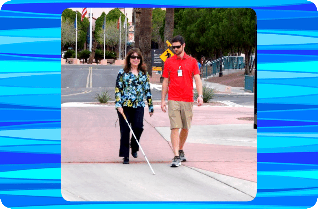 Picture of a blind woman with a cane and a sighted man walking together on a sidewalk.