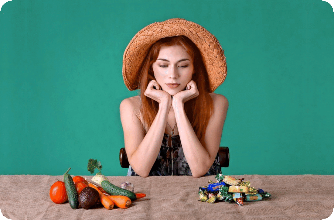 Picture of a woman seated at a table between a pile of healthy vegetables and a pile of sugary candy, trying to decide on which to choose.