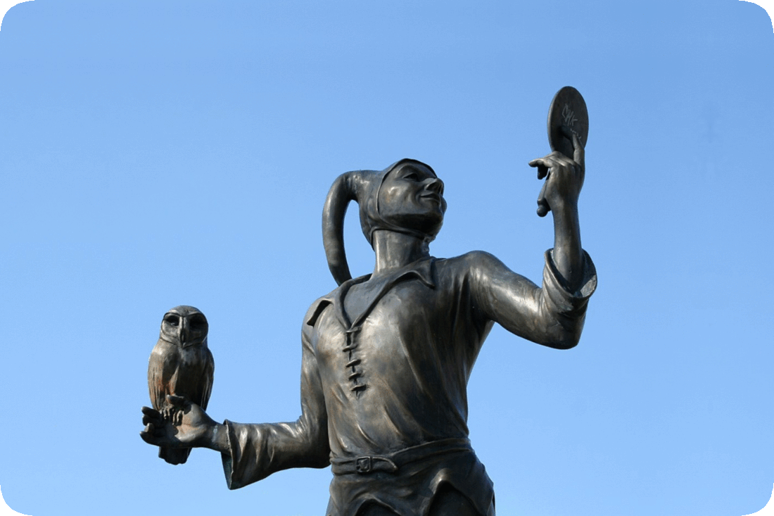 Picture of a statue of Till Eulenspiegel, a prankster character found in folk tales, in this instance holding a mirror in one hand while an owl is perched on his other hand, with a clear blue sky in the background.