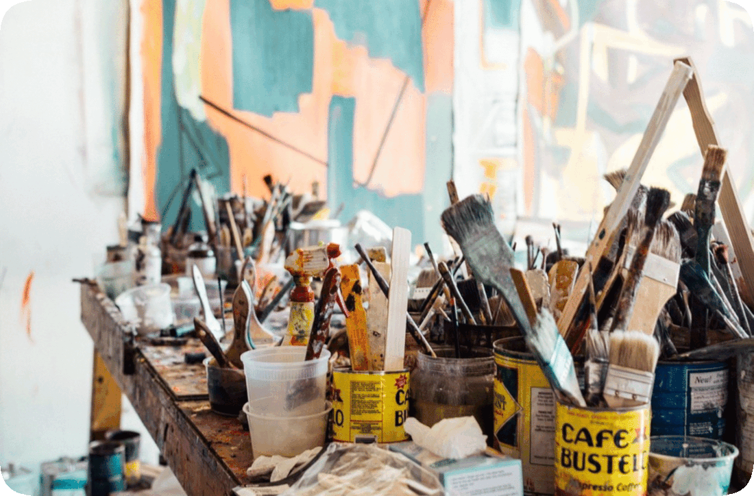 Picture of a messy table covered with containers holding paintbrushes of various sizes and stirring sticks, with splatters and spots of paint everywhere, inside an artist’s studio.