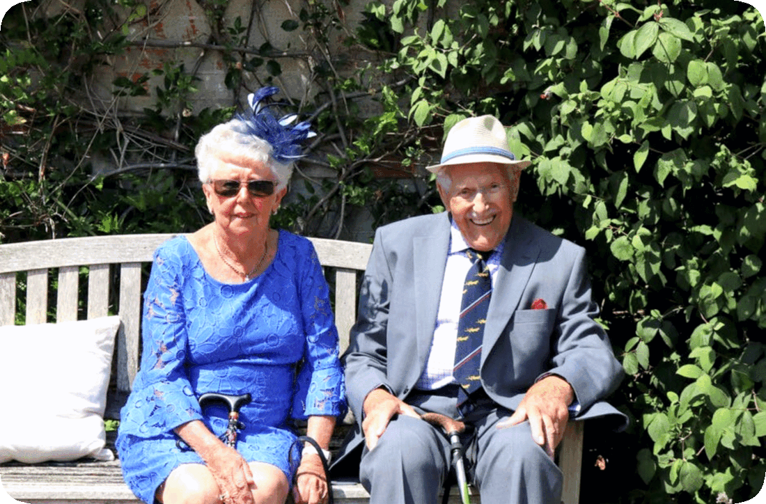 Picture of a happy, smiling older married couple outside on a bright, sunny day, seated on a wooden bench with a brick wall covered with green leafy plants behind them.