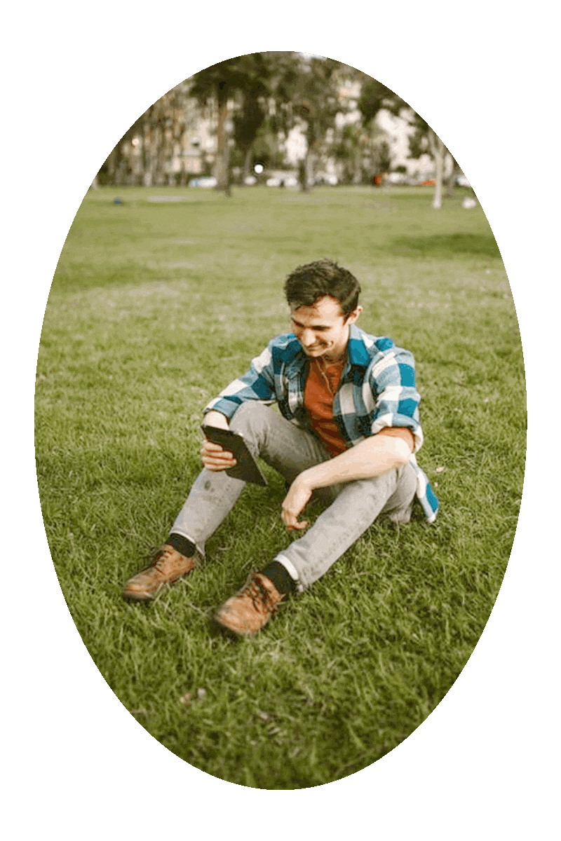 Picture of a man sitting on the ground in a green grassy area and looking at a tablet computer.