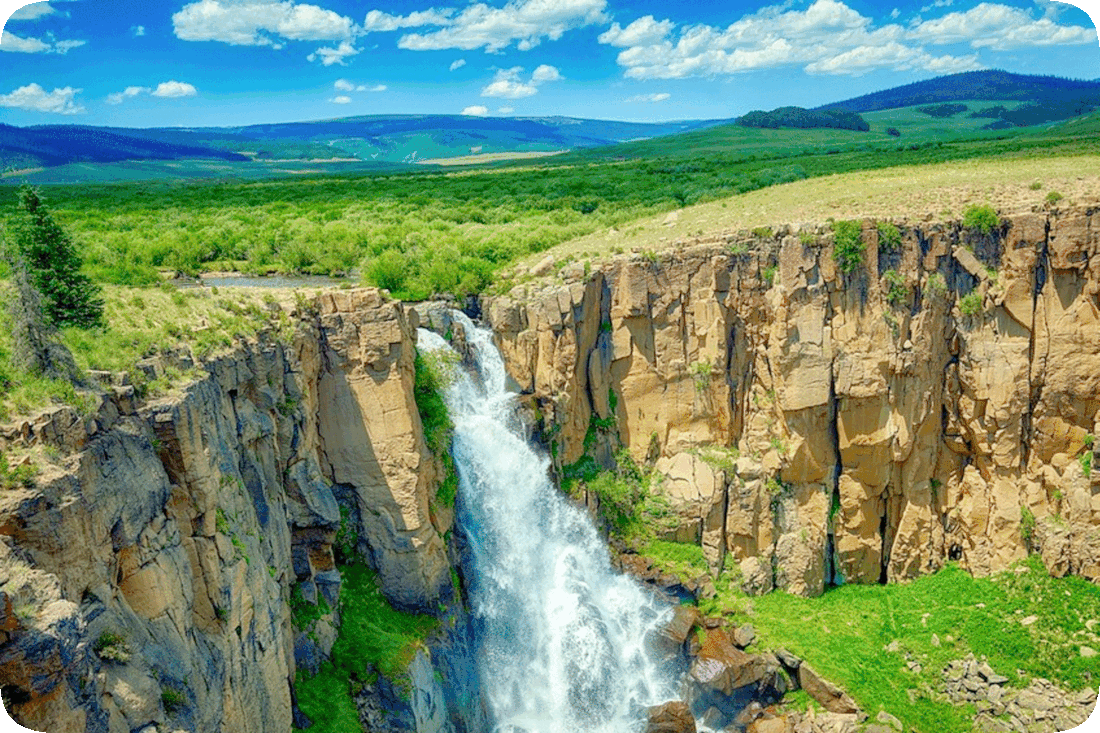 Picture of a waterfall going down over bare rocks with green grass and green bushes going into the distance at the top of the waterfall and meeting the blue sky with fluffy white clouds.