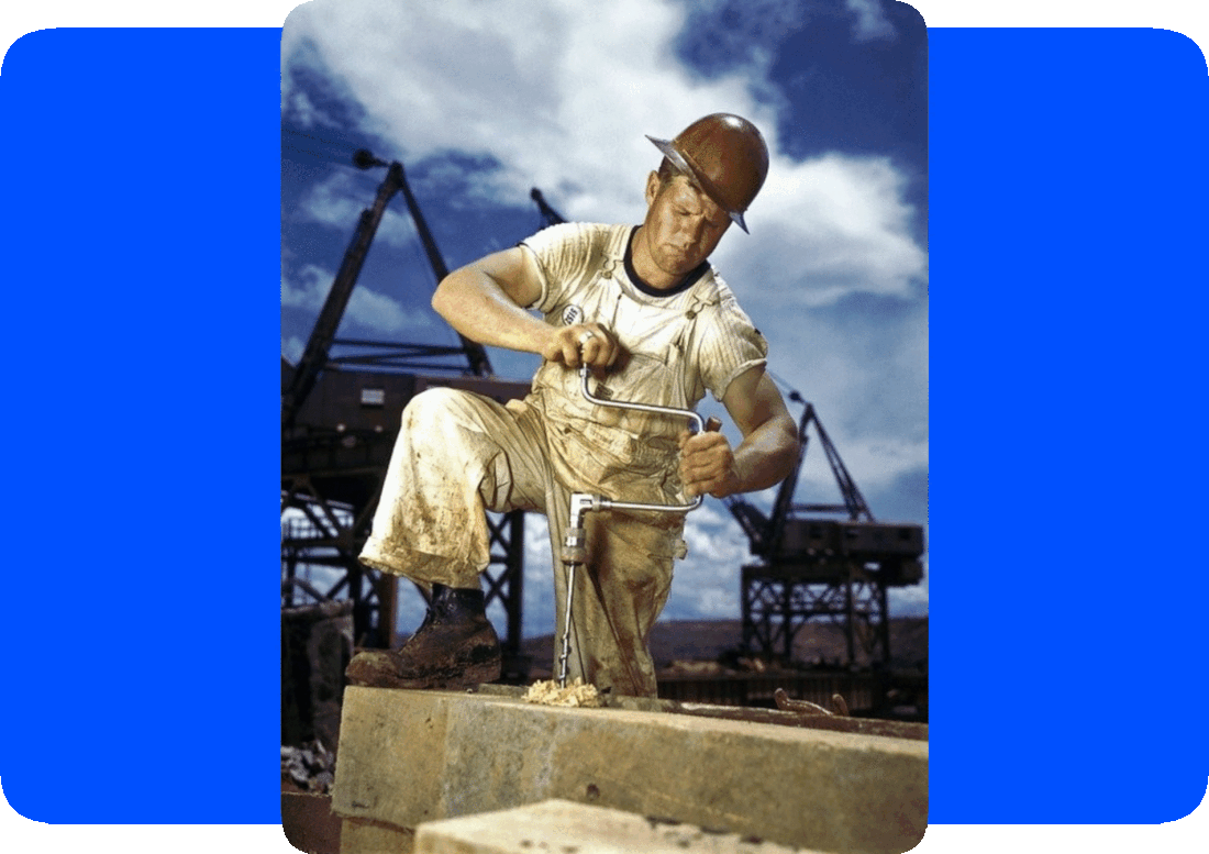 Picture of a worker wearing a hardhat at a construction site, making a hole in a wood beam using a manual hand-drill, with construction equipment, a blue sky, and fluffy white clouds in the background.