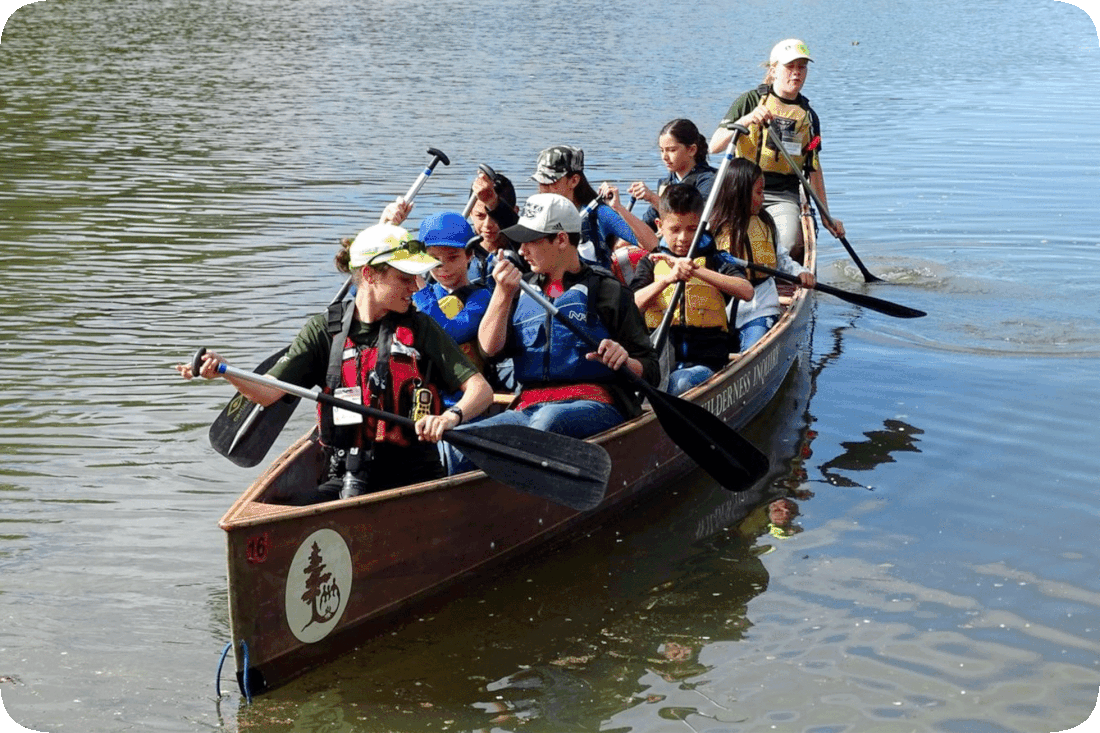 Picture of a group of nine young adults and children at a summer camp, crowded into a small canoe, each with a paddle, some of whom appear to be beginners at paddling a canoe to make it move in the water.