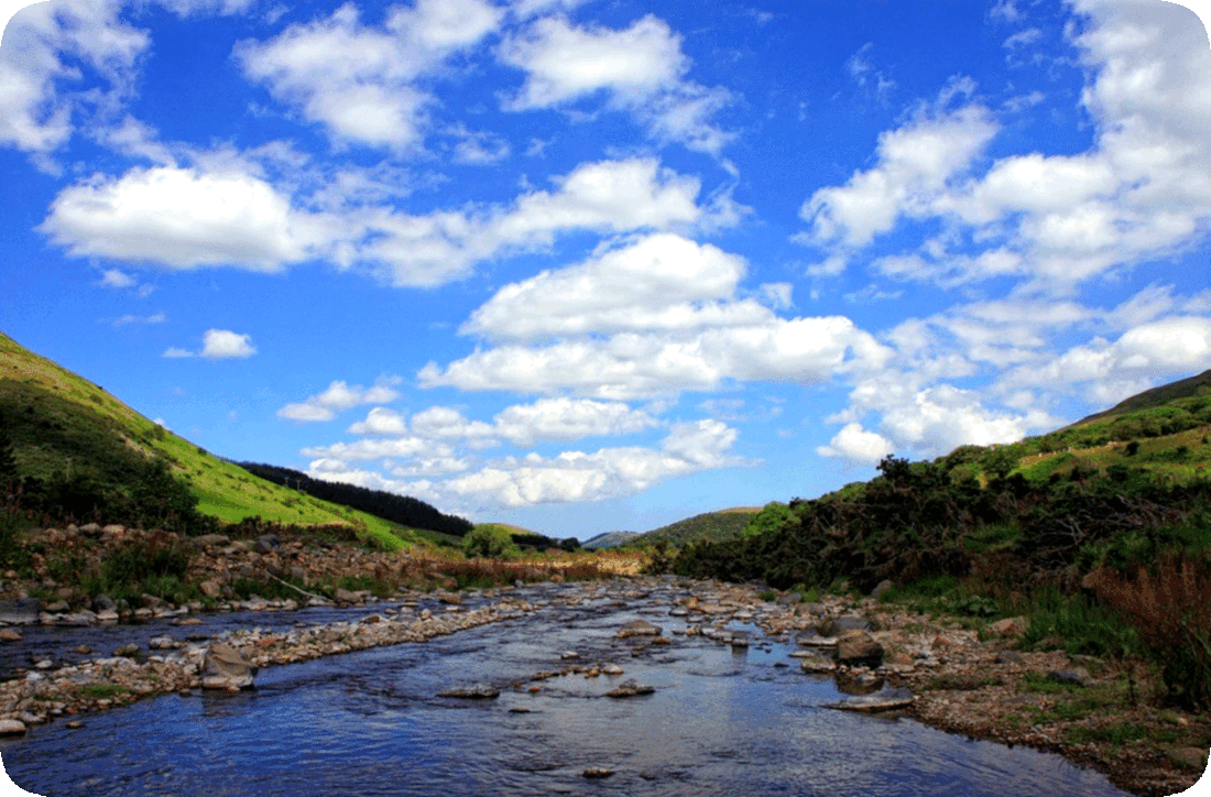 Picture of a shallow river with green grass and green leafy bushes on both of its banks, with green hill in the distance, all under a blue sky with fluffy white clouds.