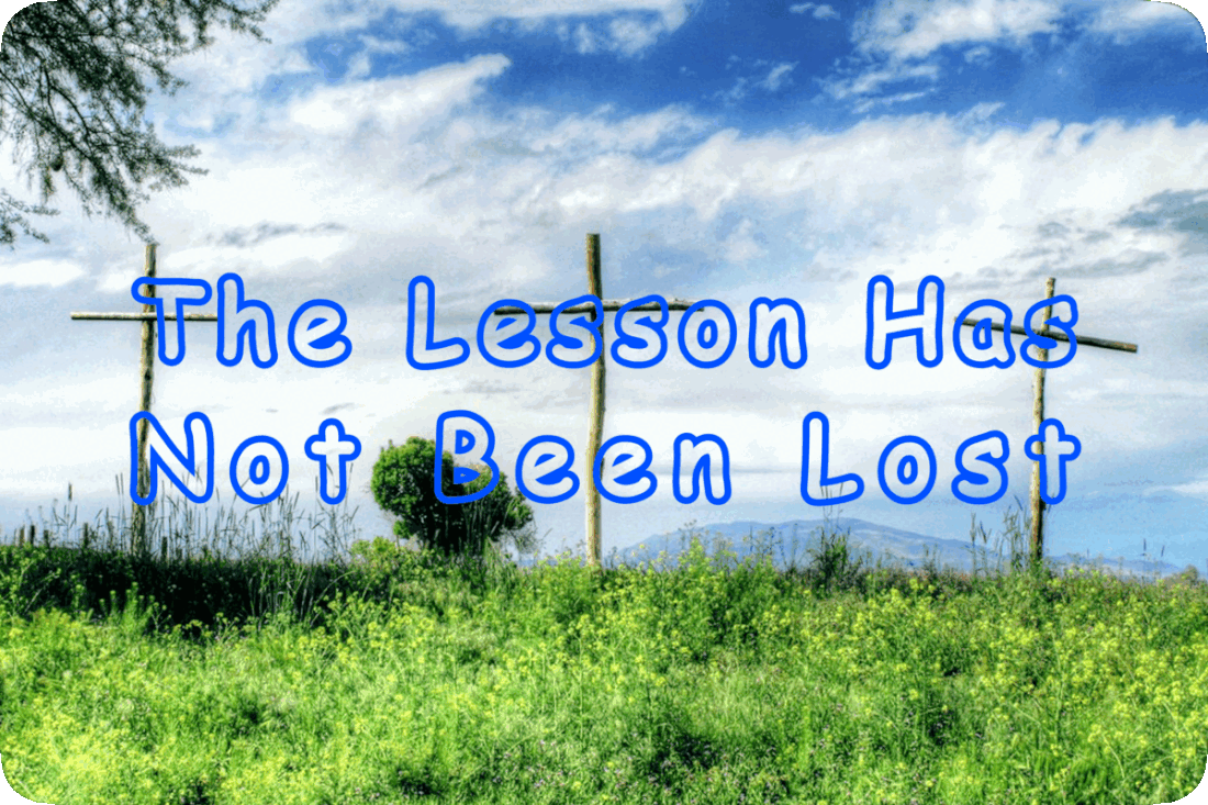 Picture of three rustic wooden crosses on a hilltop thickly covered by grasses and green plants with small yellow flowers, a blue sky with fluffy white clouds tinged with blue-gray coloration overhead, and the words, ‘The Lesson Has Not Been Forgotten.’