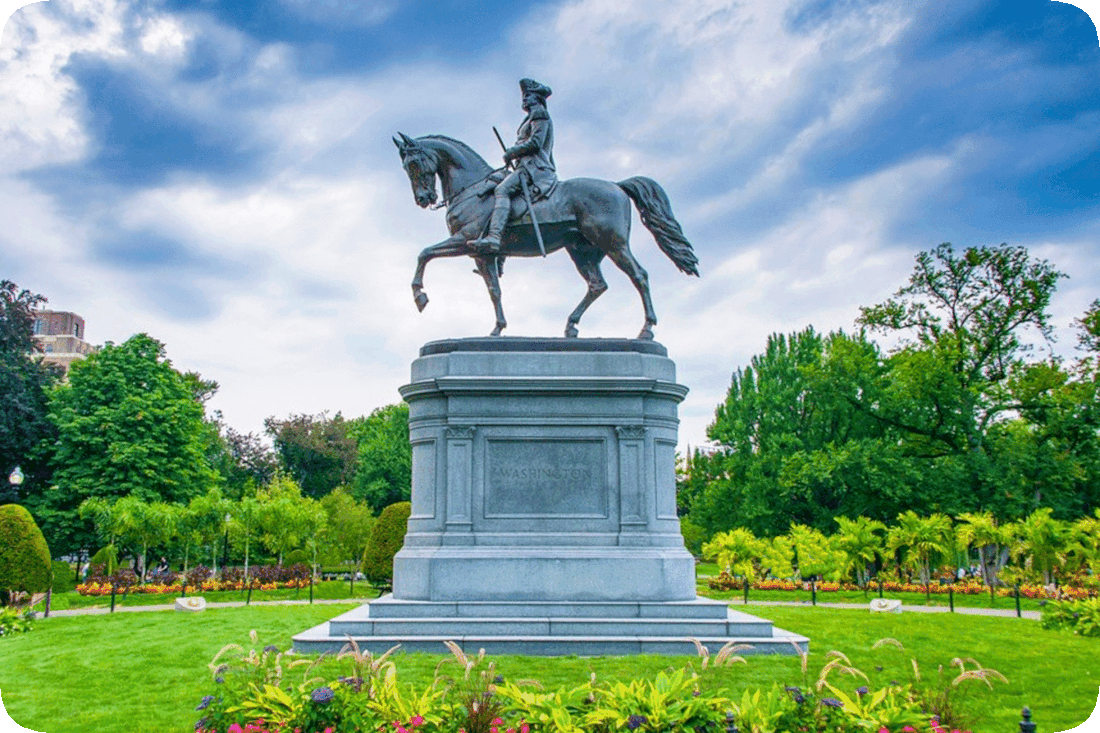 Picture of a statue of George Washington on horseback, in a park, surrounded by green grass, flowering plants, and green leafy trees.