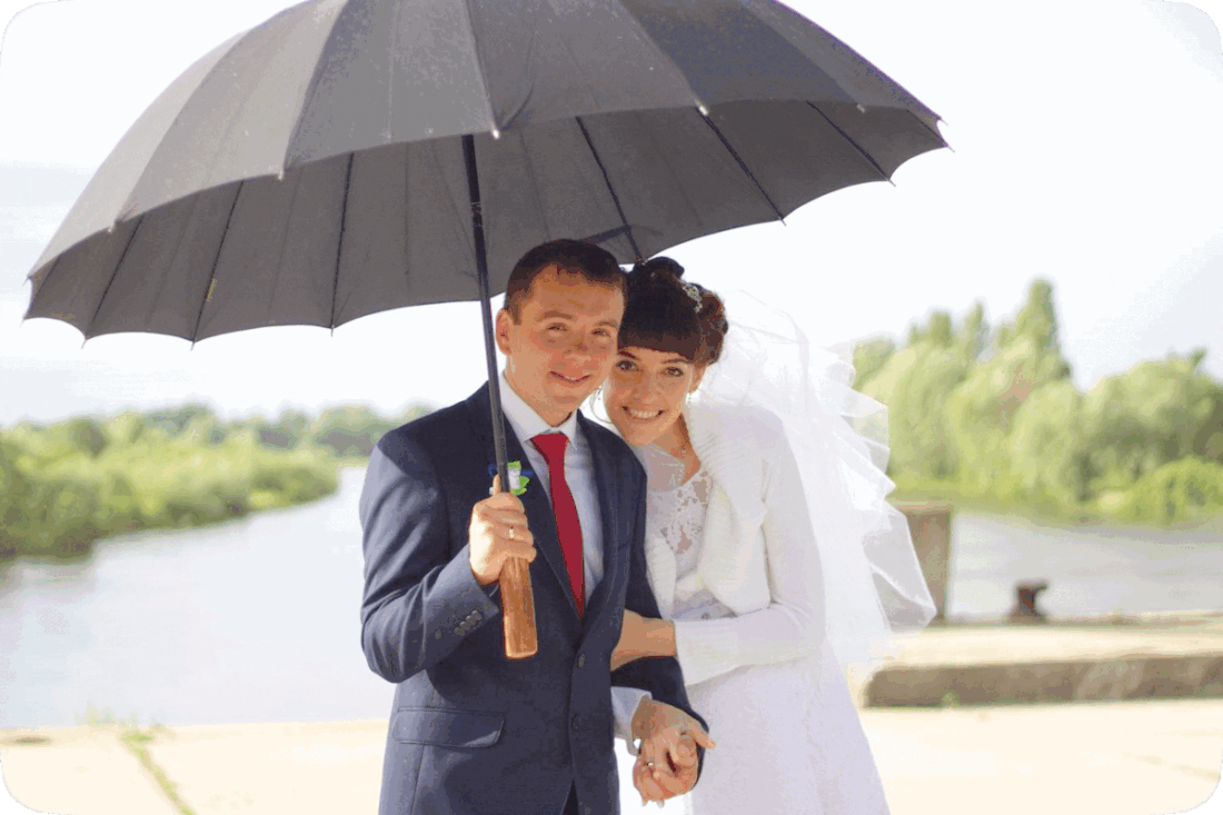Picture of a happy smiling groom and bride on their wedding day standing outside under an umbrella.