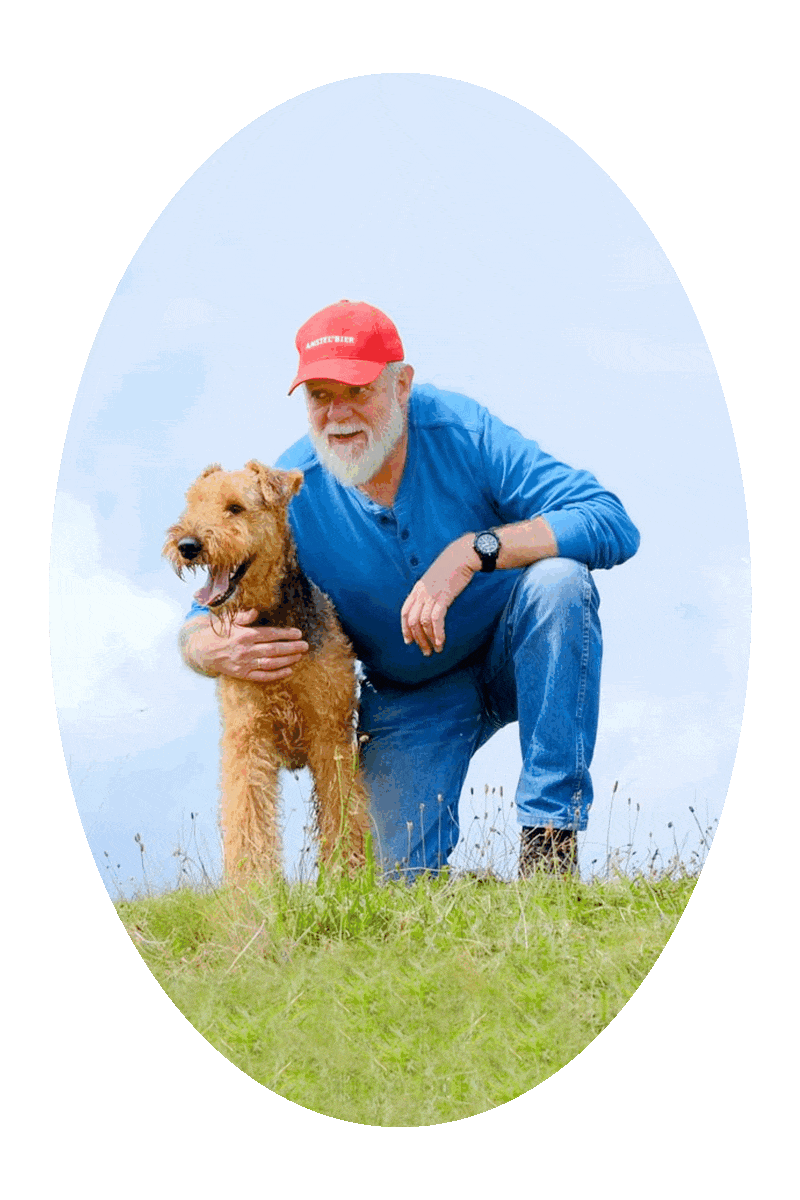 Picture of a man taking a knee next to an Airedale dog at the top of a green grassy hill, with bluish-white clouds in the background. 