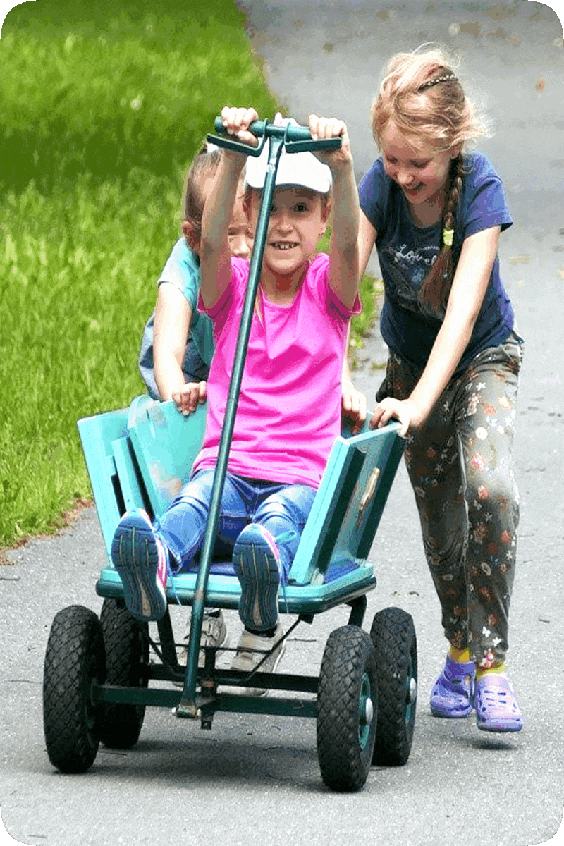 Picture of two girls pushing a toy wagon that a third girl is riding in.