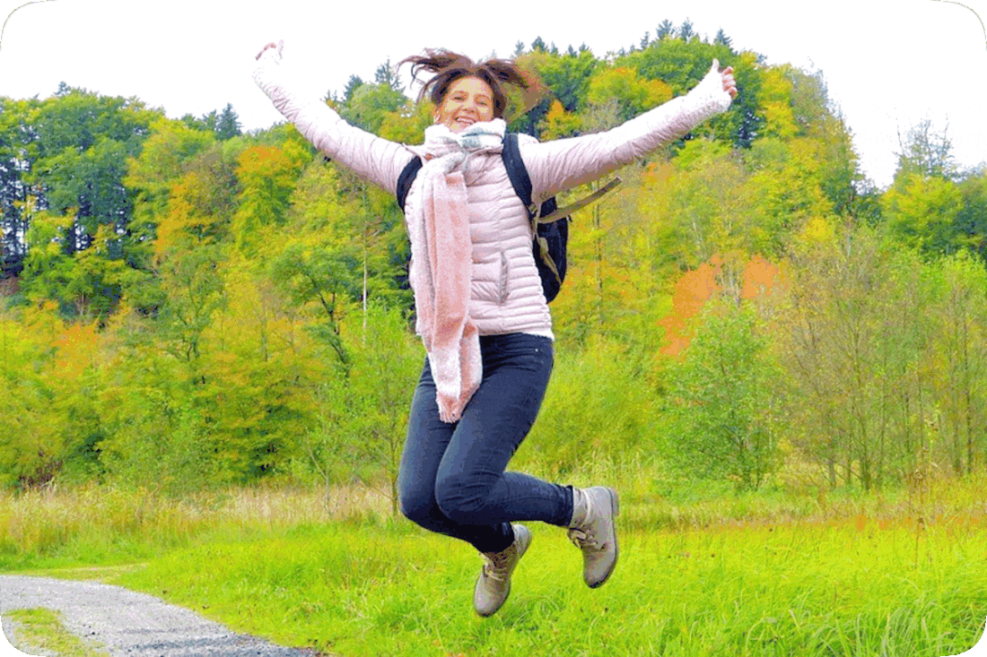 Picture of a happy, smiling woman jumping for joy outside, with green grass and green leafy trees in the background, and some of the trees turning to the oranges and yellows of early Autumn.