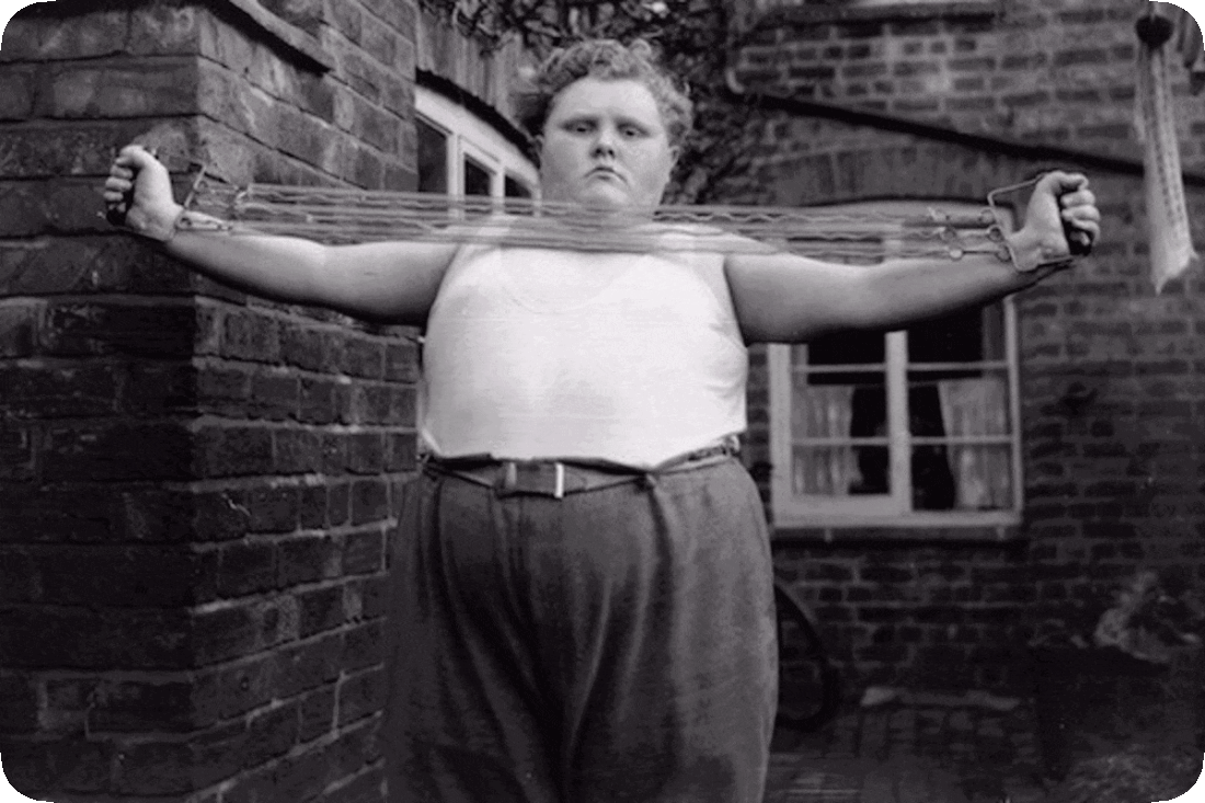 Picture of a young man standing in his back yard and using a metal-spring exercise band to build up his arm muscles.
