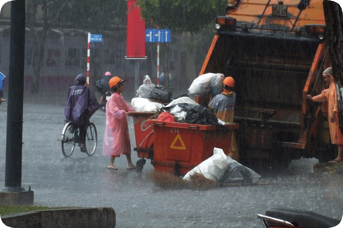 Picture of garbage collectors picking up bags of trash and putting it into a garbage truck in heavy rain with water deepening in the road around them.