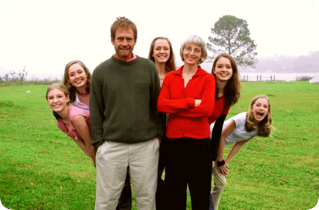 Picture of a family with a mother, father, and five children, all smiling and happy, standing outside in a grassy area with a green leafy tree and a lake in the background.