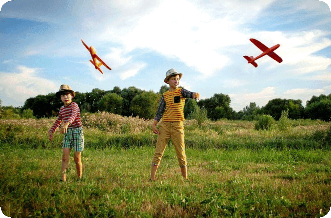 Picture of two children in a grassy field throwing gliders, or toy airplanes, into the air, with flowering plants, trees, and a sky with fluffy white clouds in the background.