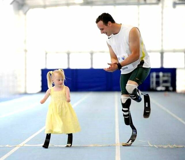 Picture of a double-leg amputee man and a quadruple-legs-and-arms amputee toddler girl both wearing prosthetic legs and crossing the finish line of an indoor race track.