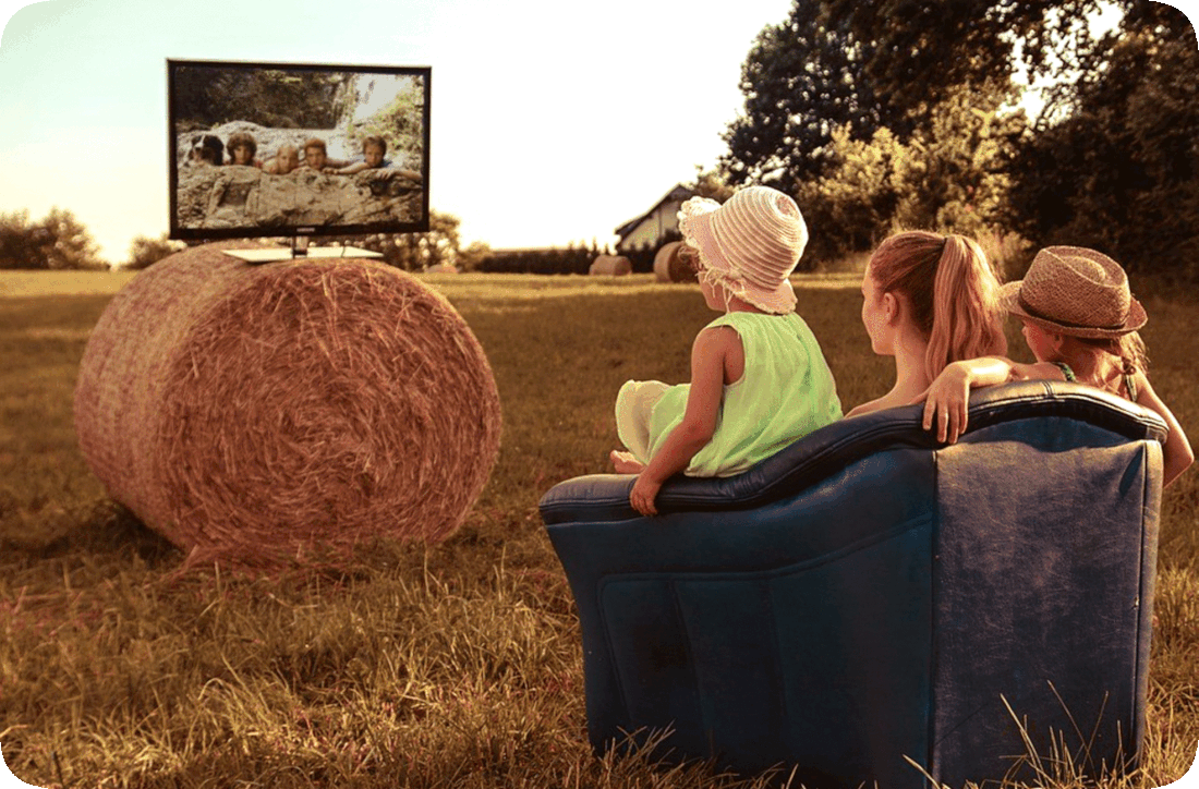 Picture of three people sitting in a chair outside in a field, watching a television that is on top of a bale of hay.