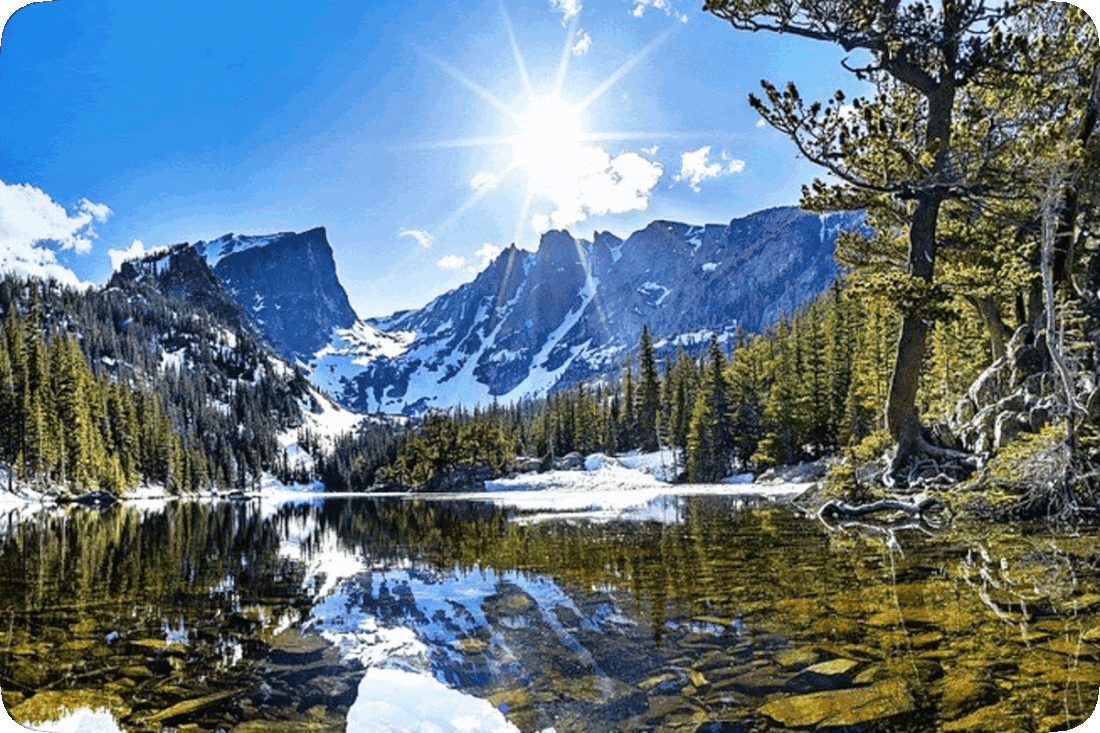 Picture of a crystal clear mountain lake surrounded by evergreen trees, with snow-covered mountains at a distance, and the Sun brightly shining above, in a blue sky with scattered fluffy white clouds.
