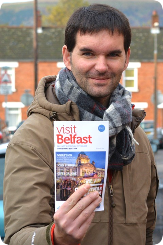 Picture of a happy smiling Irishman holding up a travel brochure with the printed words, 'Visit Belfast.'