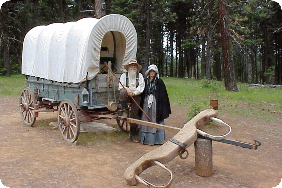 Picture of an American pioneer couple, a man and a woman, standing next to a covered wagon, with tall evergreen trees in the background.