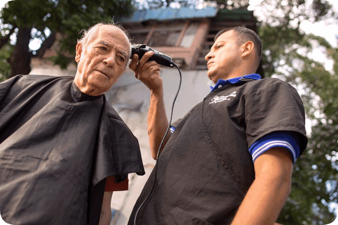 Picture of a barber using electric shears to cut a man’s hair outdoors, with green leafy trees in the background.